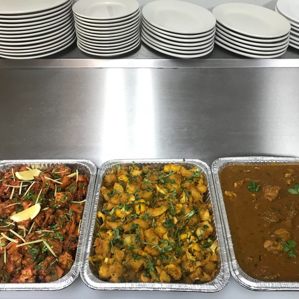 Three foil trays of Indian dishes, including a curry, on a metal counter with stacked plates.
