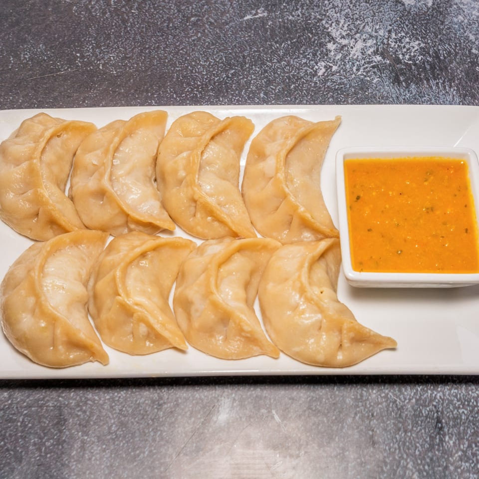 A dozen steamed Nepalese momos, or dumplings, with a side of orange dipping sauce on a white plate.
