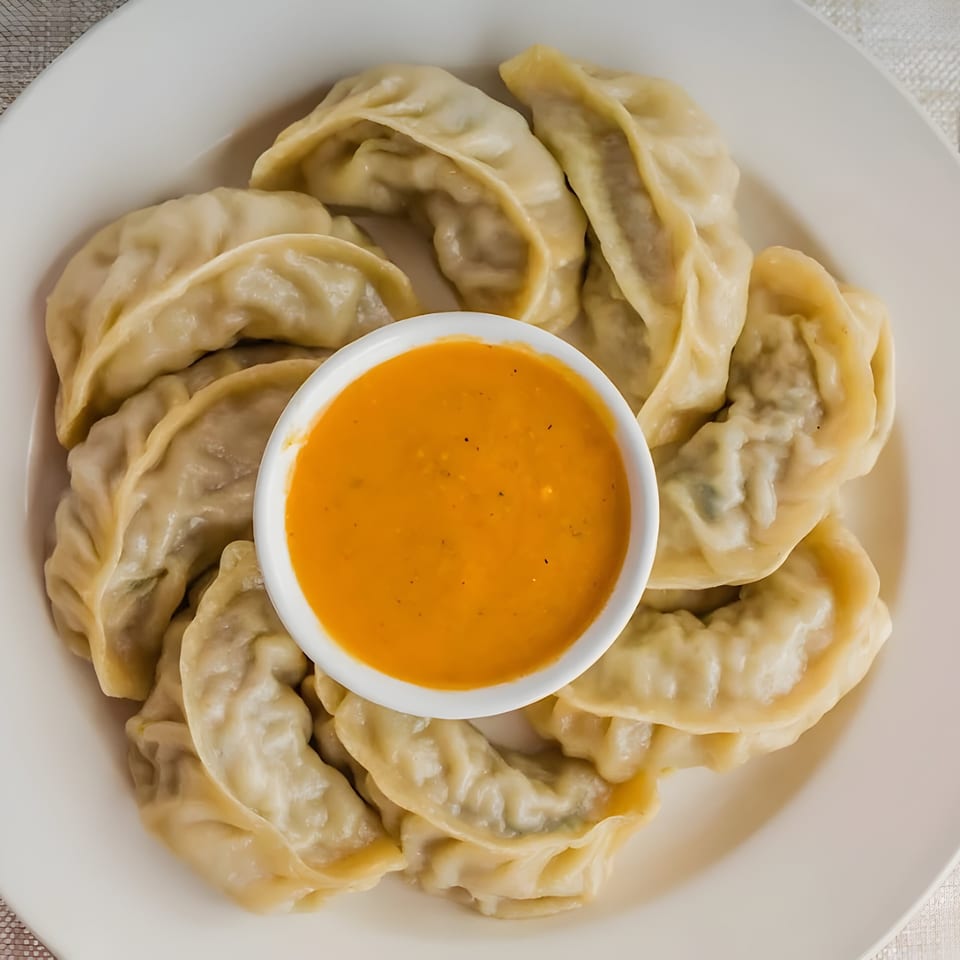 A plate of steamed Nepalese momo dumplings with a small bowl of orange dipping sauce.